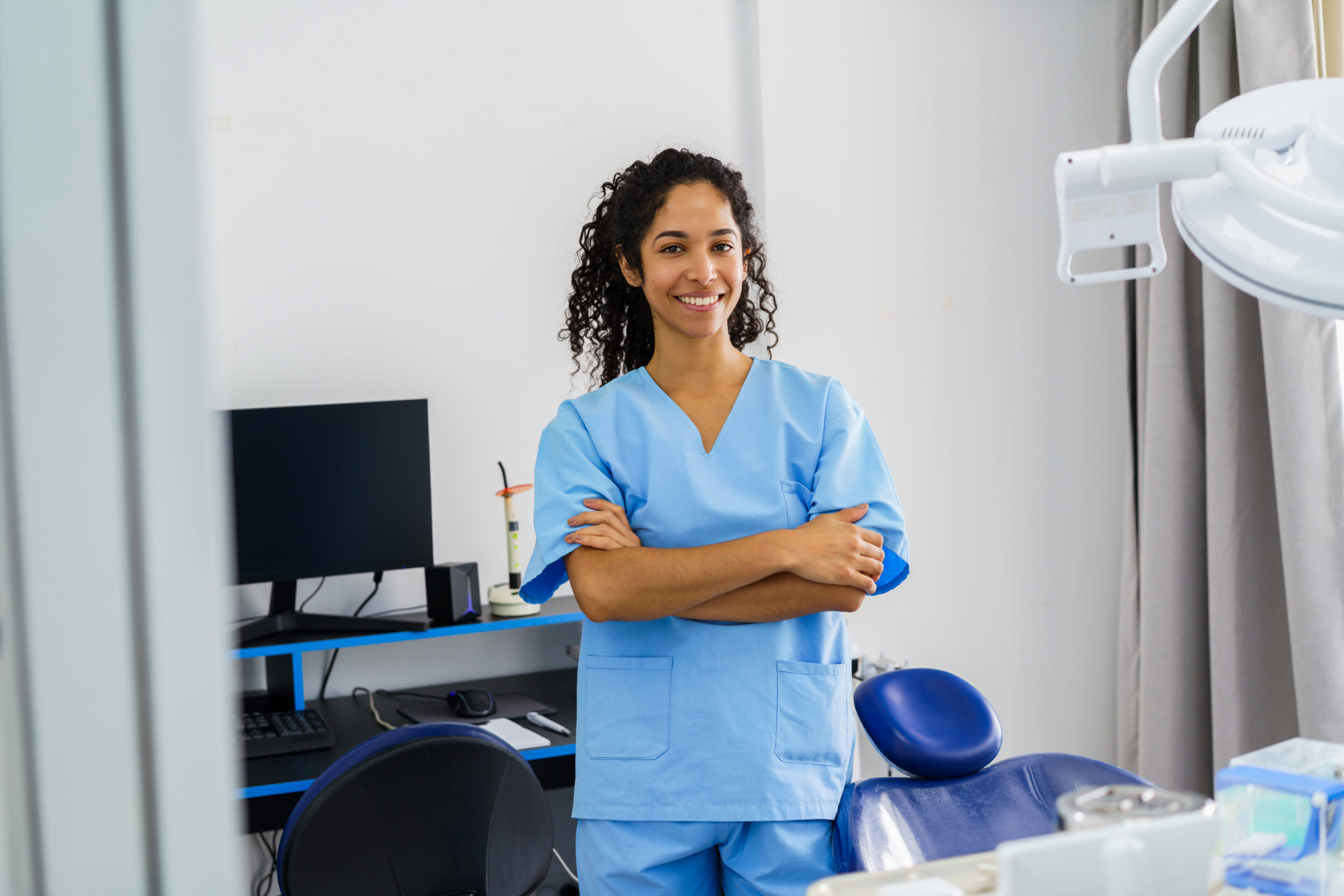 A smiling healthcare professional in light blue scrubs stands with arms crossed inside a dental clinic. Modern equipment and a computer visible in the background convey professionalism, care, and accessible patient-focused dentistry.