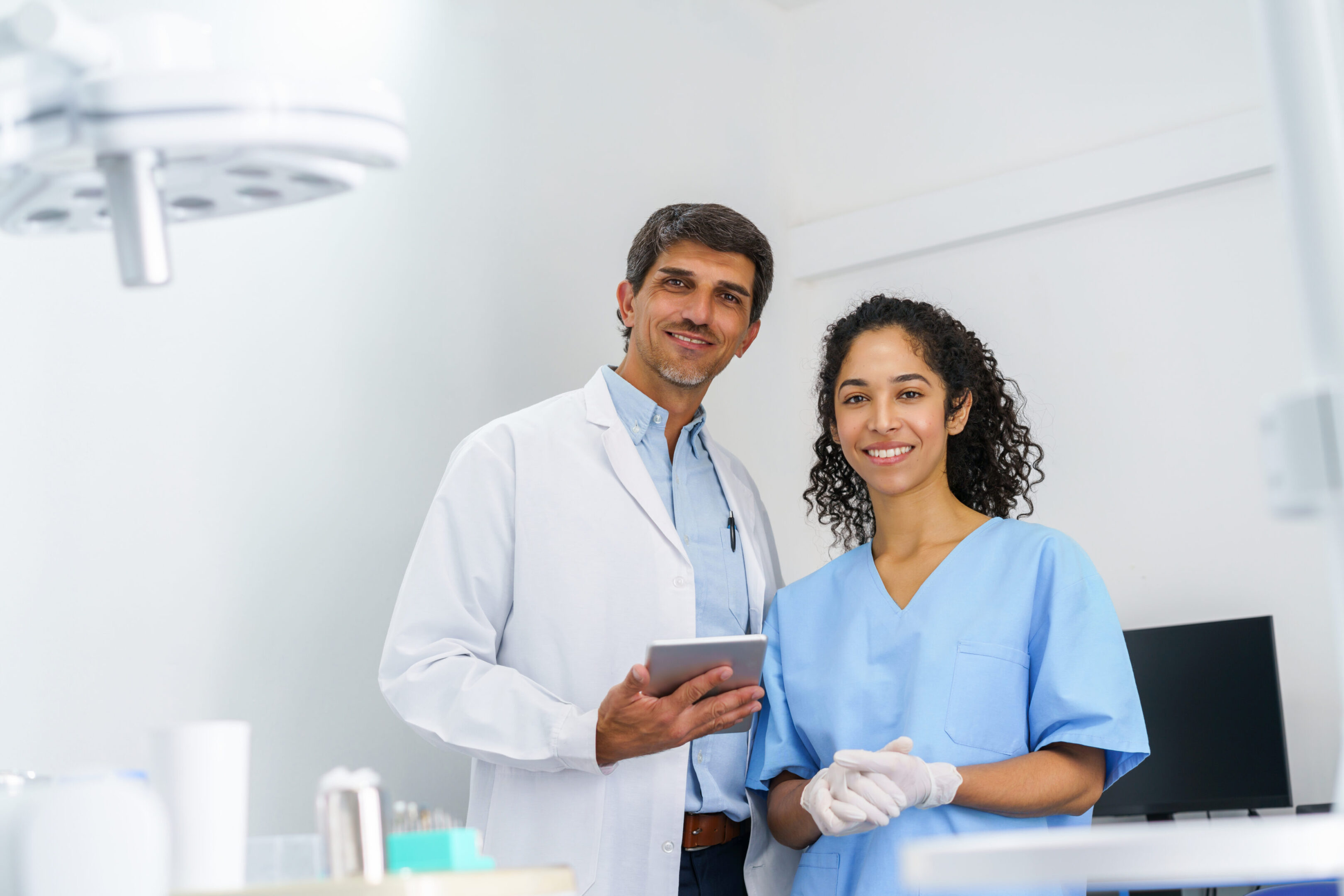 A friendly dental team stands together in a clean, modern clinic. One dentist holds a tablet while a nurse in scrubs smiles, conveying teamwork, care, and professional patient service.