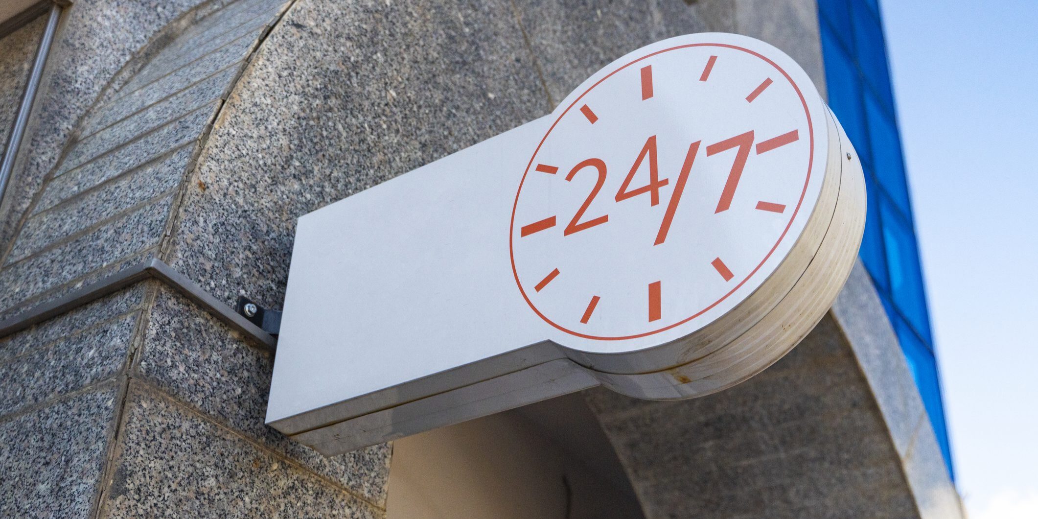 A white banner with the image of a clock signifying the round-the-clock operation of the institution against the background of a blue sky. twenty-four by seven round-the-clock work signage